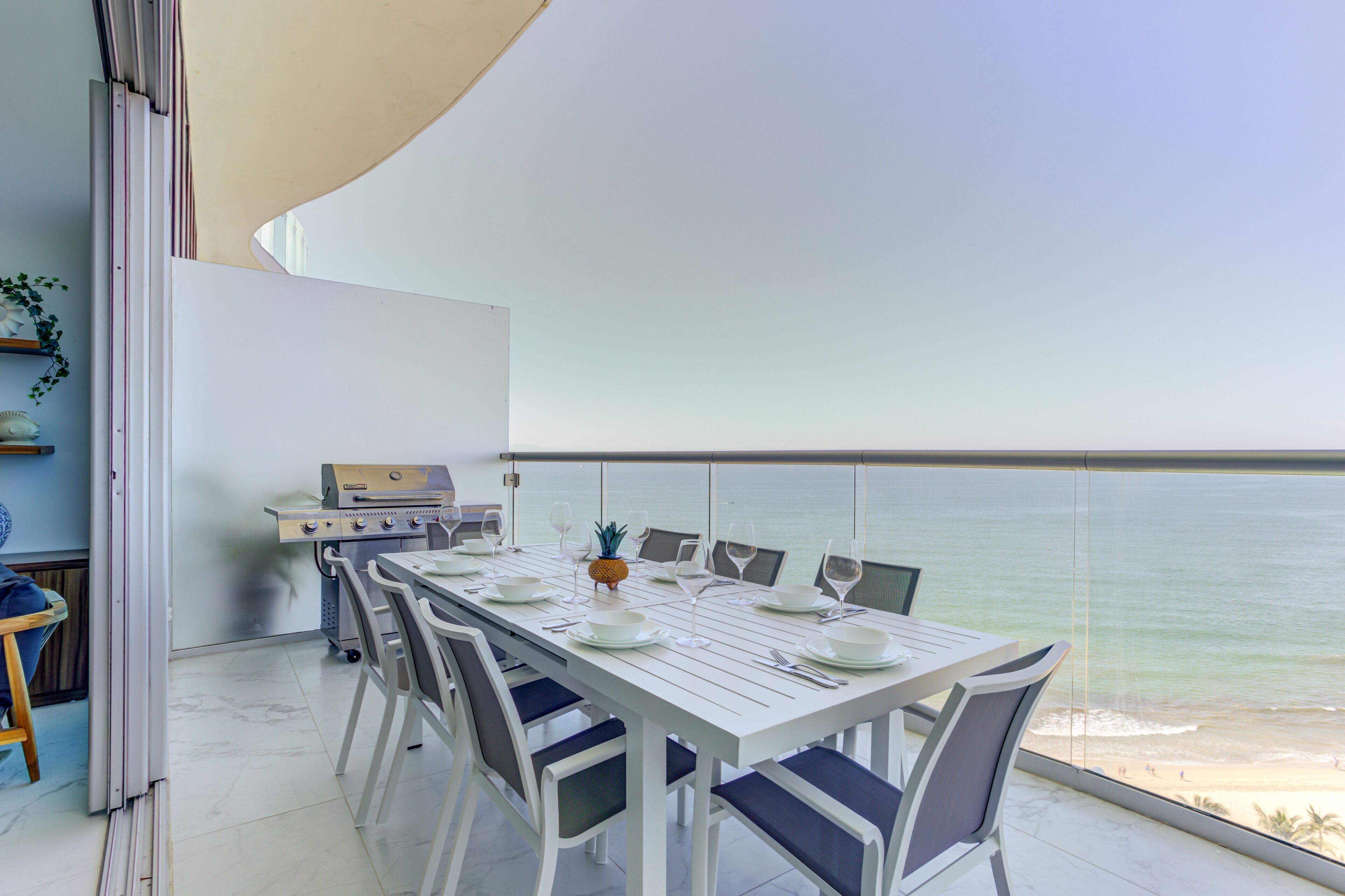 A dining area with a white table set for six, overlooking a beach.