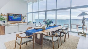 A spacious living area with a large window overlooking the sea, a wooden dining table, and a comfortable seating arrangement. - Marítima Beachfront Playa Flamingos (Nuevo Nayarit)