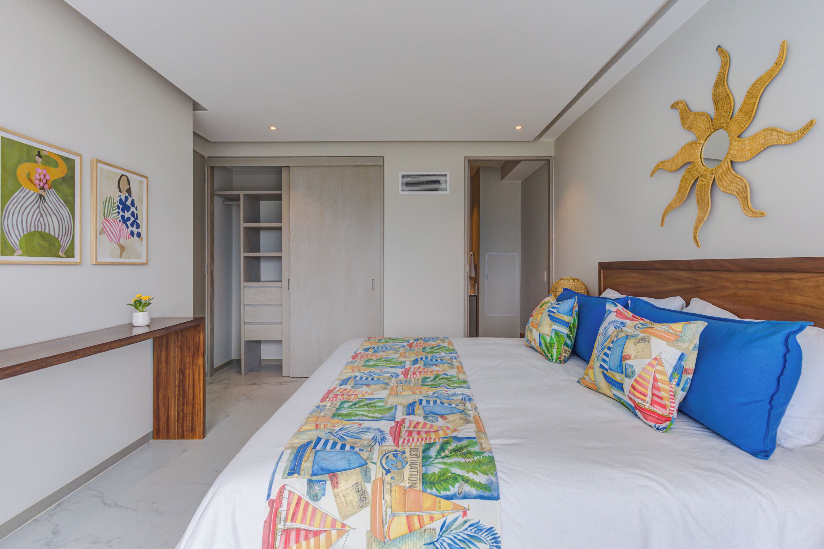 Une chambre avec un grand lit, des coussins colorés, une tête de lit en bois, une décoration murale représentant un soleil et deux œuvres d’art encadrées accrochées au mur.