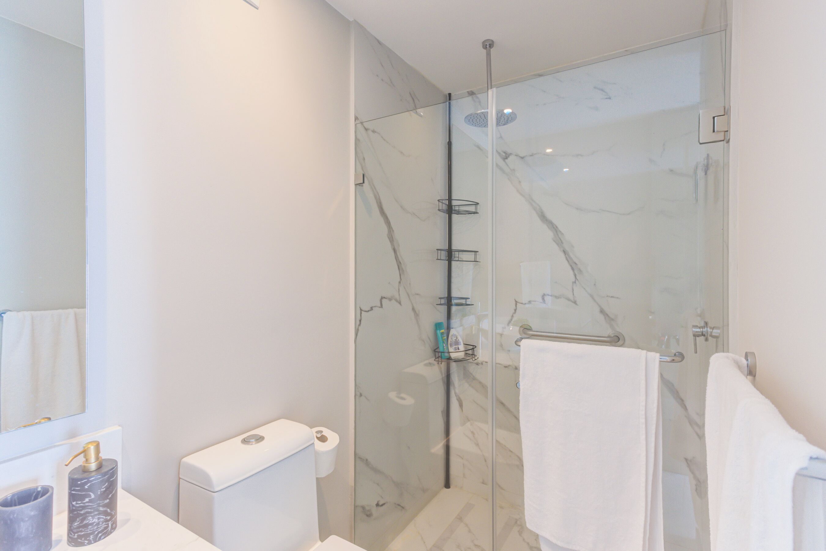 A modern bathroom with a glass-enclosed shower, marble walls, a toilet, and a sink with a marble soap dispenser.