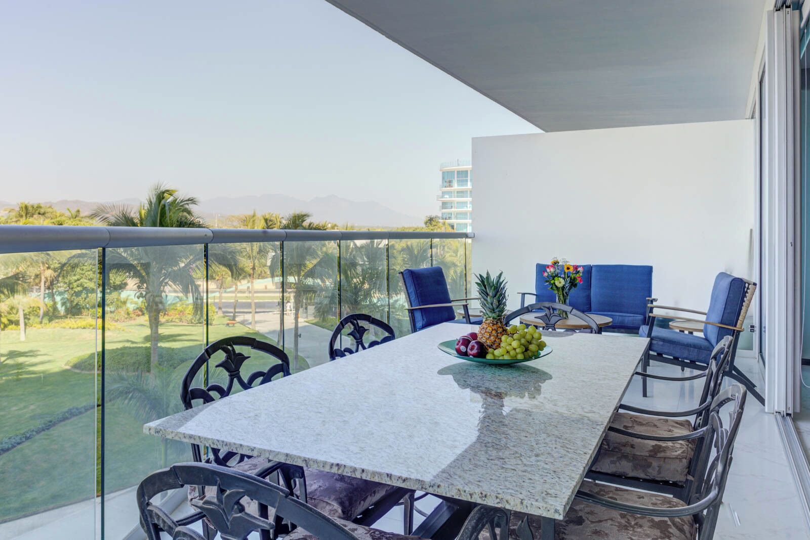 A balcony with a table set for dining, featuring a fruit bowl, and blue upholstered chairs.
