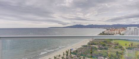 A coastal view with a beach, palm trees, and a hotel in the background.