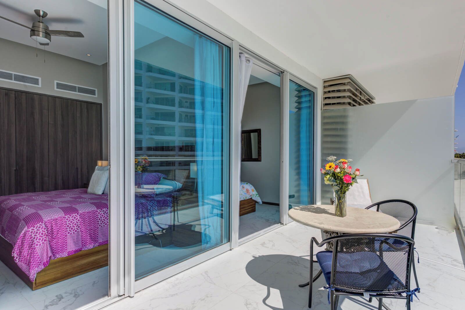 A modern room with a bed, a table with chairs, and a balcony with a view of buildings.