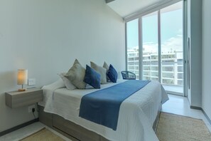 A modern bedroom with a large bed, a nightstand, a lamp, and a view of a building through a large window. - Marítima Beachfront Playa Flamingos (Nuevo Nayarit)