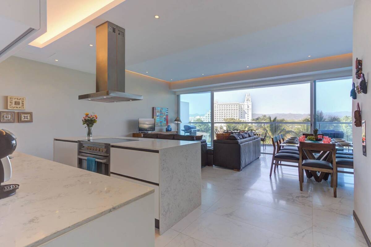 Cocina moderna con una isla grande, comedor con vistas y sala de estar con sofá y televisor.