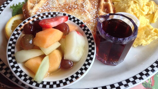 A plate with a fruit bowl, a glass of dark beverage, scrambled eggs, and a waffle. - Hotel Luxe Stay (New Delhi)