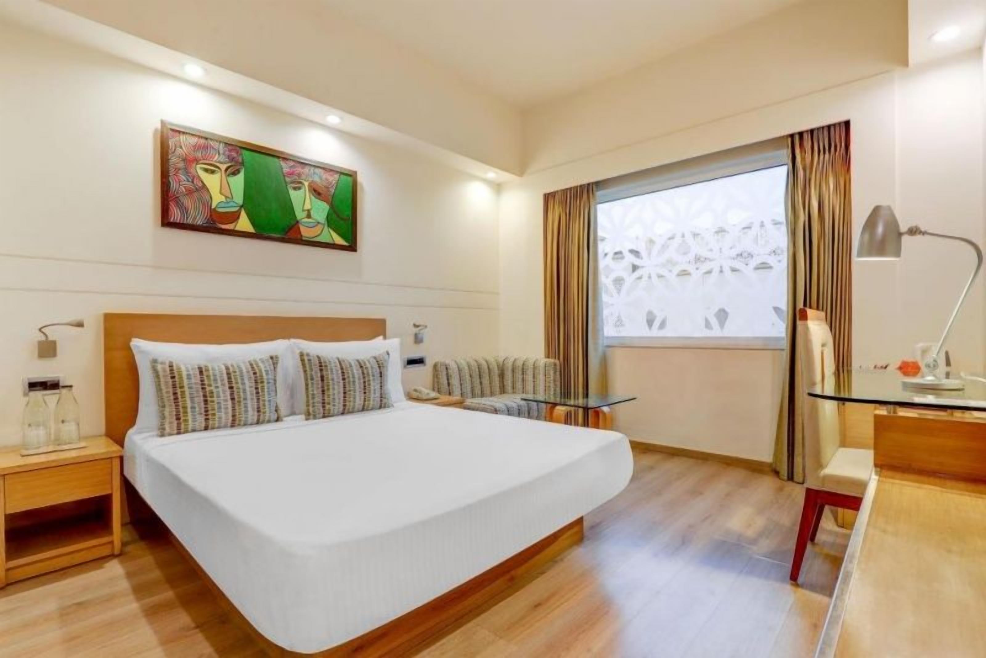 A hotel room with a large bed, a bedside table with a lamp, a window with curtains, and a painting on the wall.