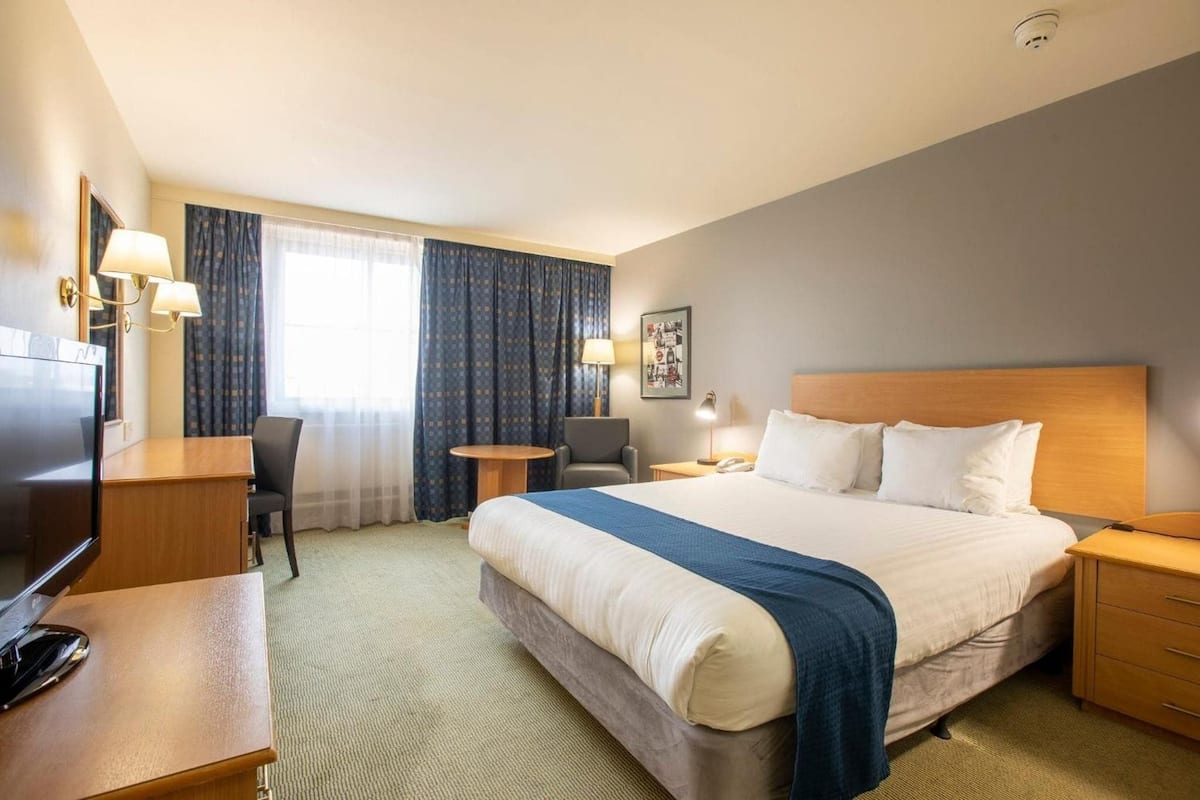 A hotel room with a large bed, a desk, a chair, a TV, and a window with curtains.