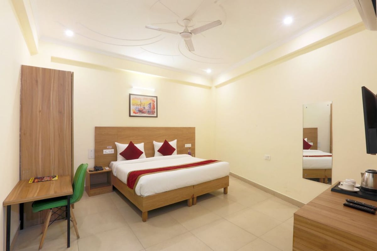 A hotel room with a bed, a desk, a chair, a ceiling fan, a painting, a mirror, and a television.