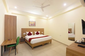 A hotel room with a bed, a desk, a chair, a ceiling fan, a painting, a mirror, and a television. - Hotel Luxe Stay (New Delhi)