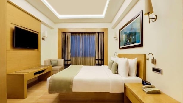 Une chambre d’hôtel moderne avec un grand lit, un téléviseur fixé au mur, une table de chevet avec une lampe et une fenêtre avec des rideaux.