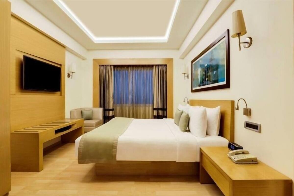Una habitación de hotel moderna con una cama grande, un televisor montado en la pared, una mesita de noche con lámpara y una ventana con cortinas.