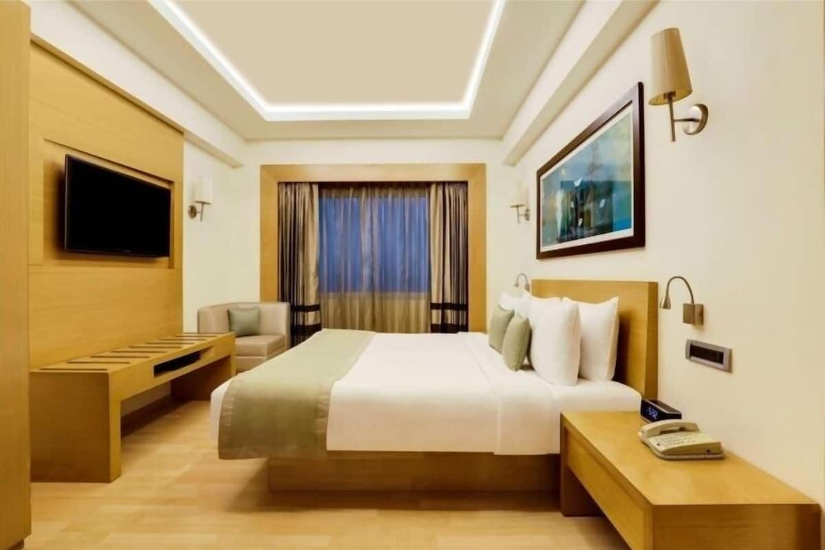 A modern hotel room with a large bed, a TV mounted on the wall, a bedside table with a lamp, and a window with curtains.