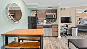Cocina compacta con refrigerador, microondas y tostadora. Hay una mesa de comedor con sillas, un pequeño espacio de oficina con escritorio y silla, y un televisor.