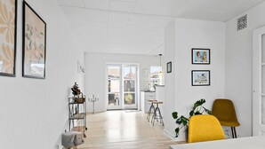 A bright, open-plan living space with a wooden floor, a dining area with yellow chairs, and a kitchen visible through an open door.
