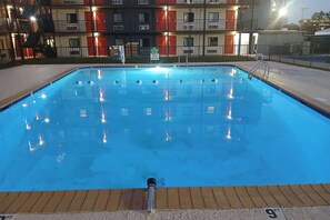 A rectangular swimming pool with a metal ladder and a safety sign. The pool is surrounded by a fence and a building with balconies in the background. - Ramada By Wyndham Kenner/New Orleans Airport South (Kenner)