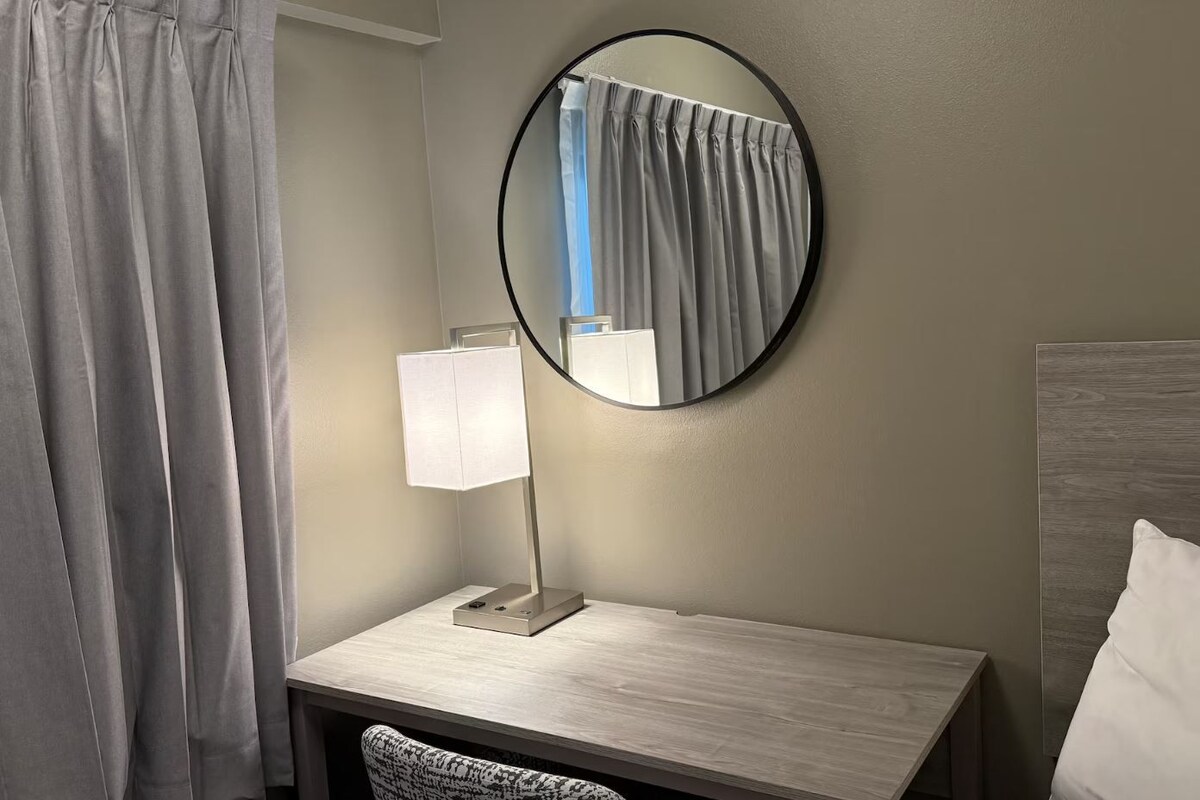 A hotel room with a wooden desk, a round mirror, a white lamp, and a grey curtain.