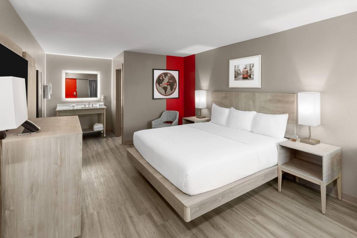 A modern hotel room with a large bed, a desk, a chair, a vanity with a mirror, and artwork on the walls.