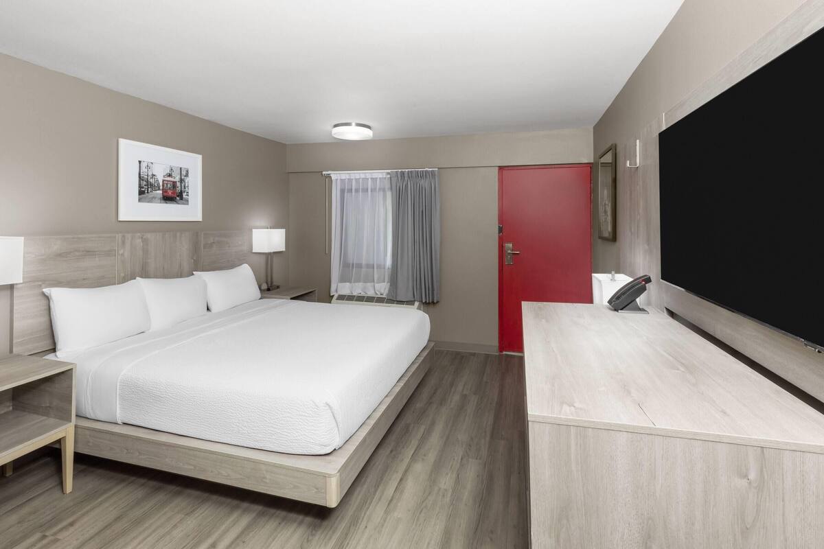 A hotel room with a large bed, a desk, a chair, a TV, a window with curtains, and a door.