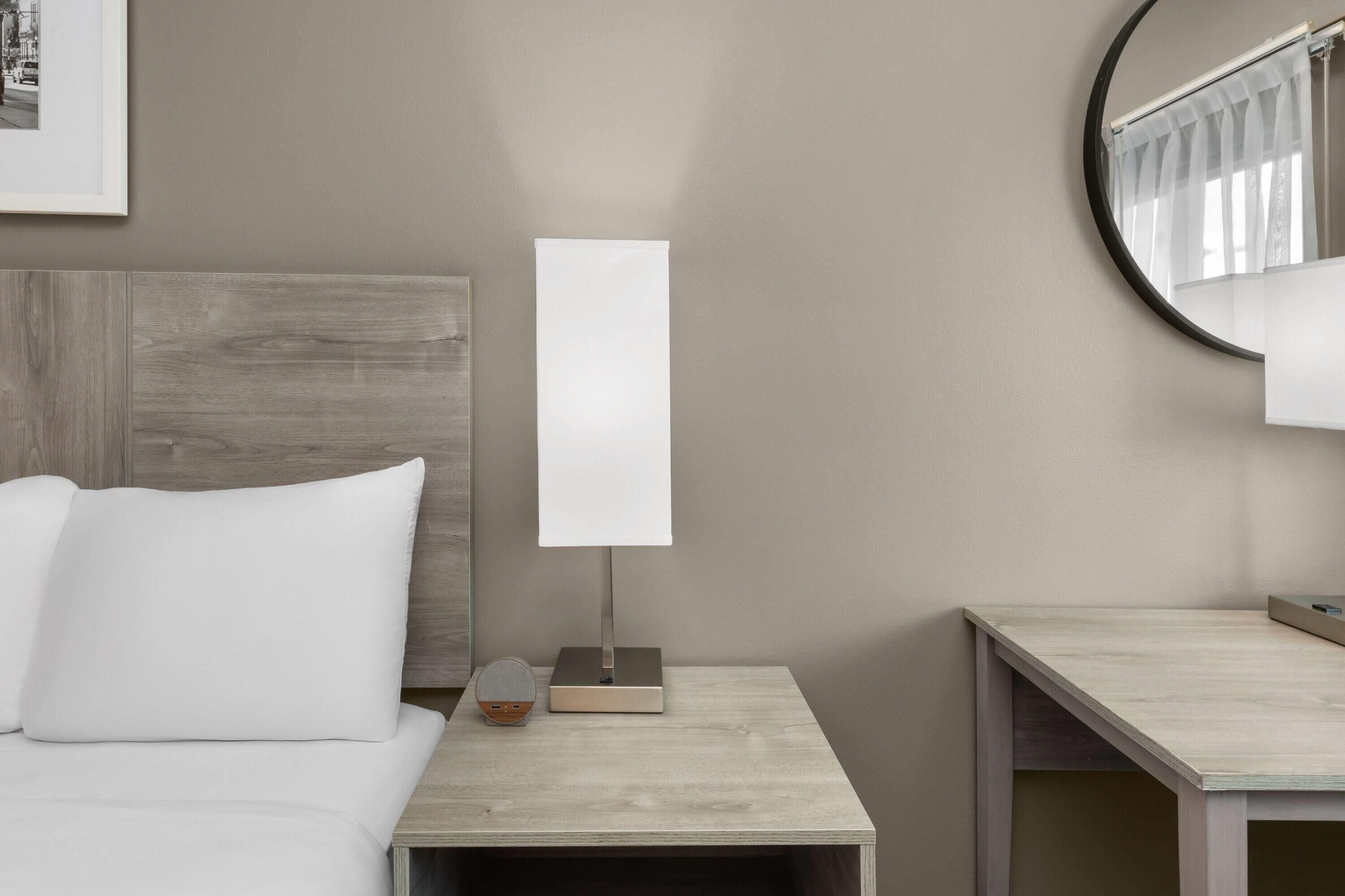 A modern hotel room with a wooden headboard, a bedside table with a lamp, and a small side table.