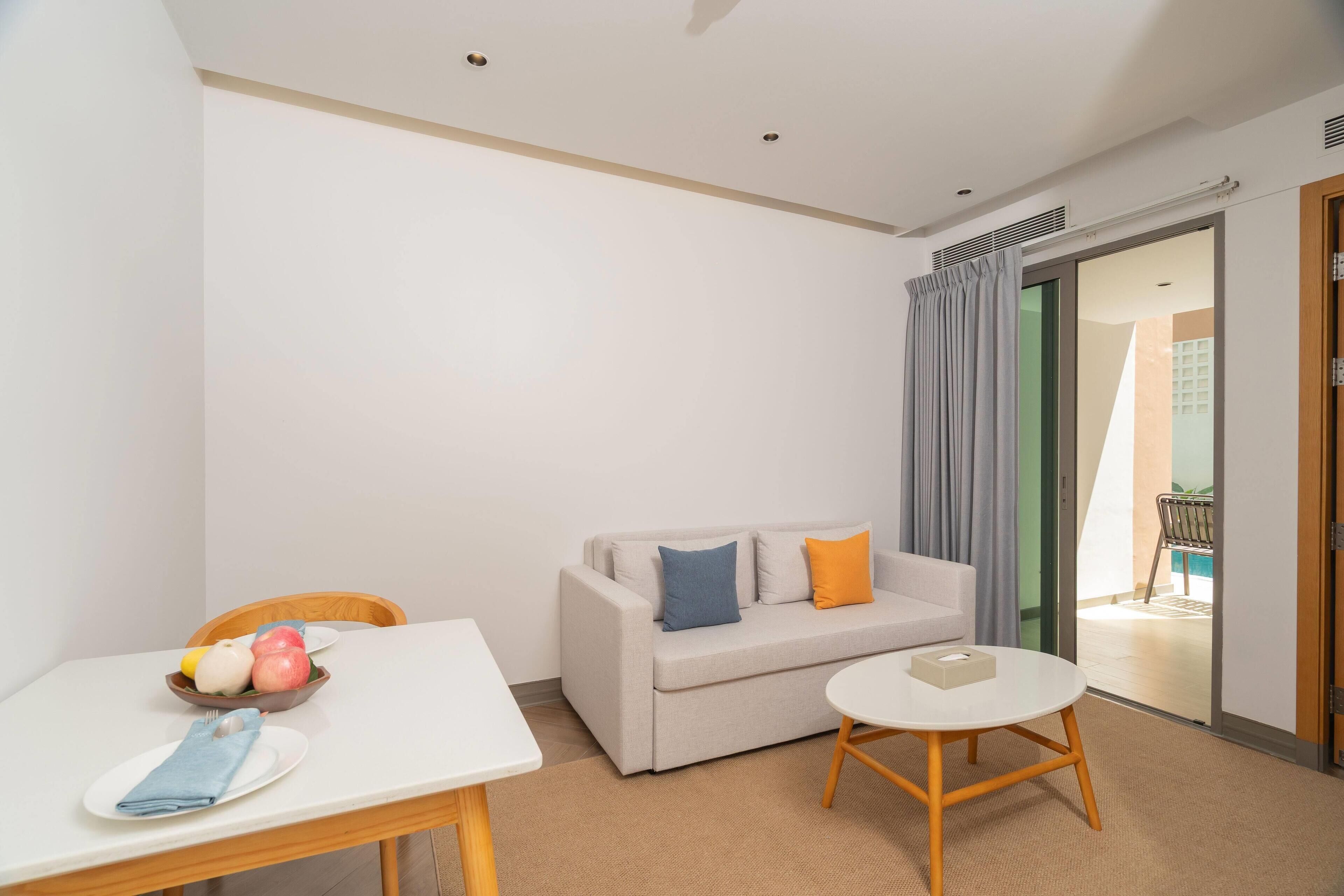 A modern hotel room with a white table, a beige sofa with cushions, a small round table, and a sliding glass door leading to a balcony.