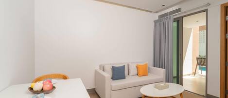 A modern hotel room with a white table, a beige sofa with cushions, a small round table, and a sliding glass door leading to a balcony.