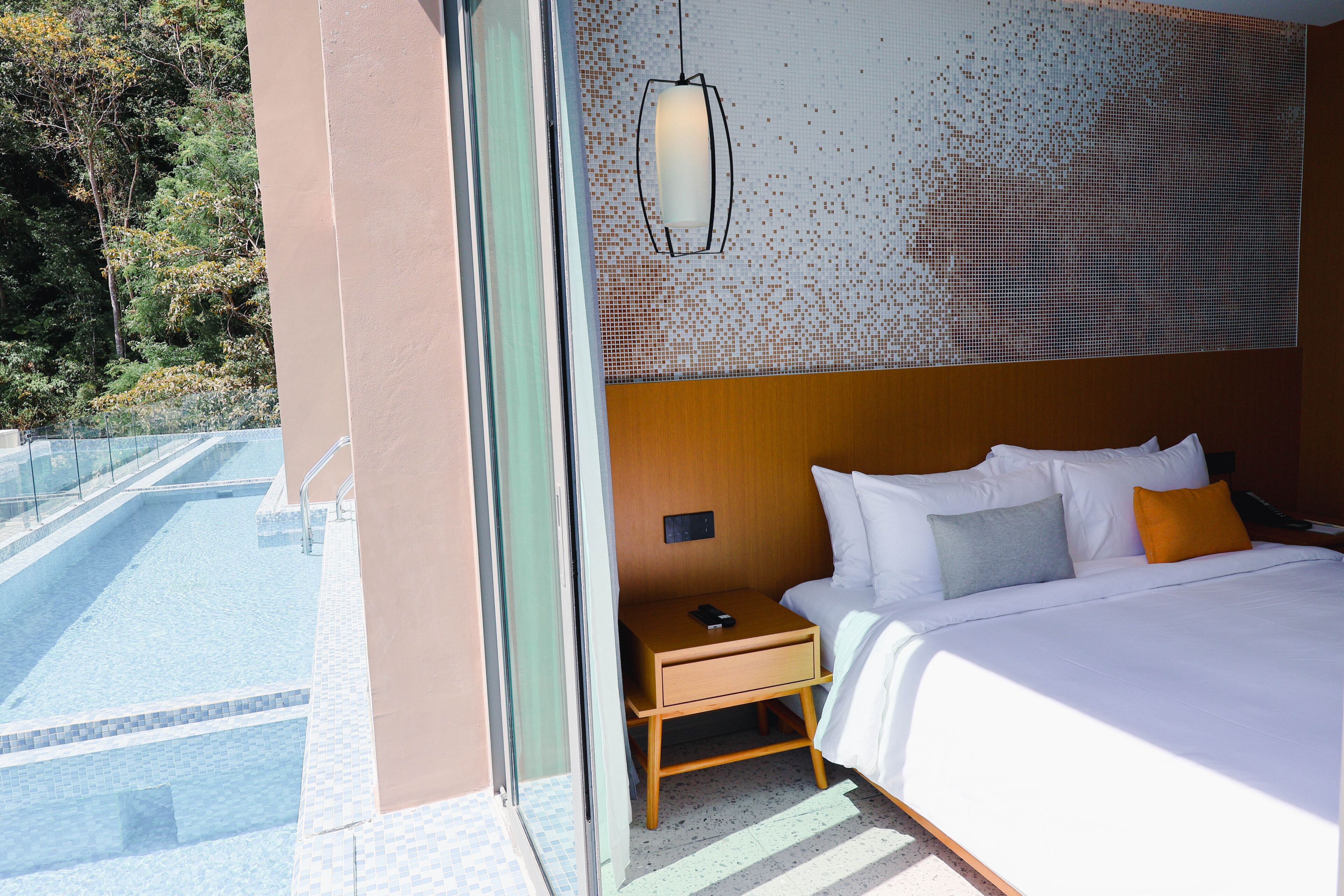 A hotel room with a bed, bedside table, and a view of a pool.