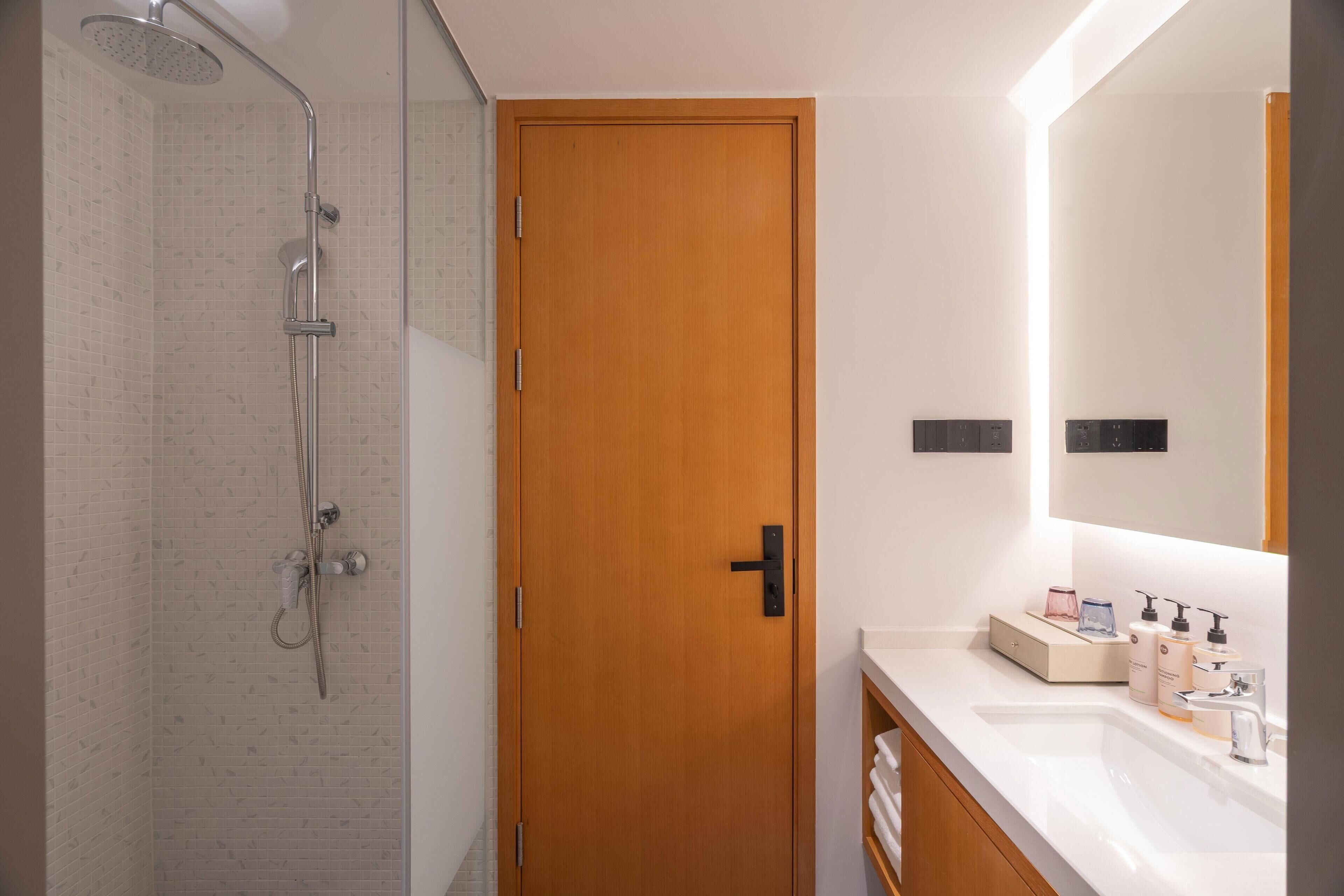 A bathroom with a shower, a wooden door, a white sink with toiletries, and a mirror above the sink.