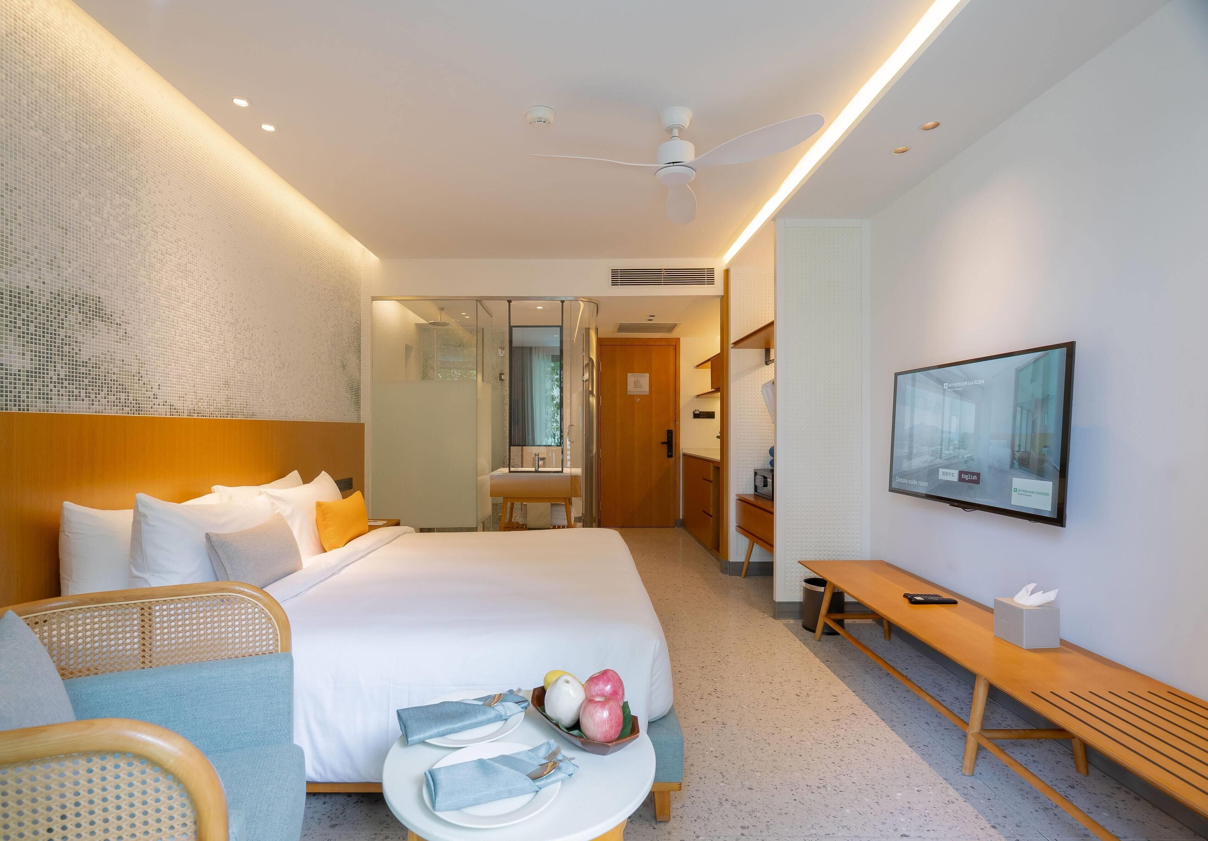 Habitación de hotel con cama, zona de estar con un banco, una mesita con un frutero, un televisor en la pared y un ventilador de techo.