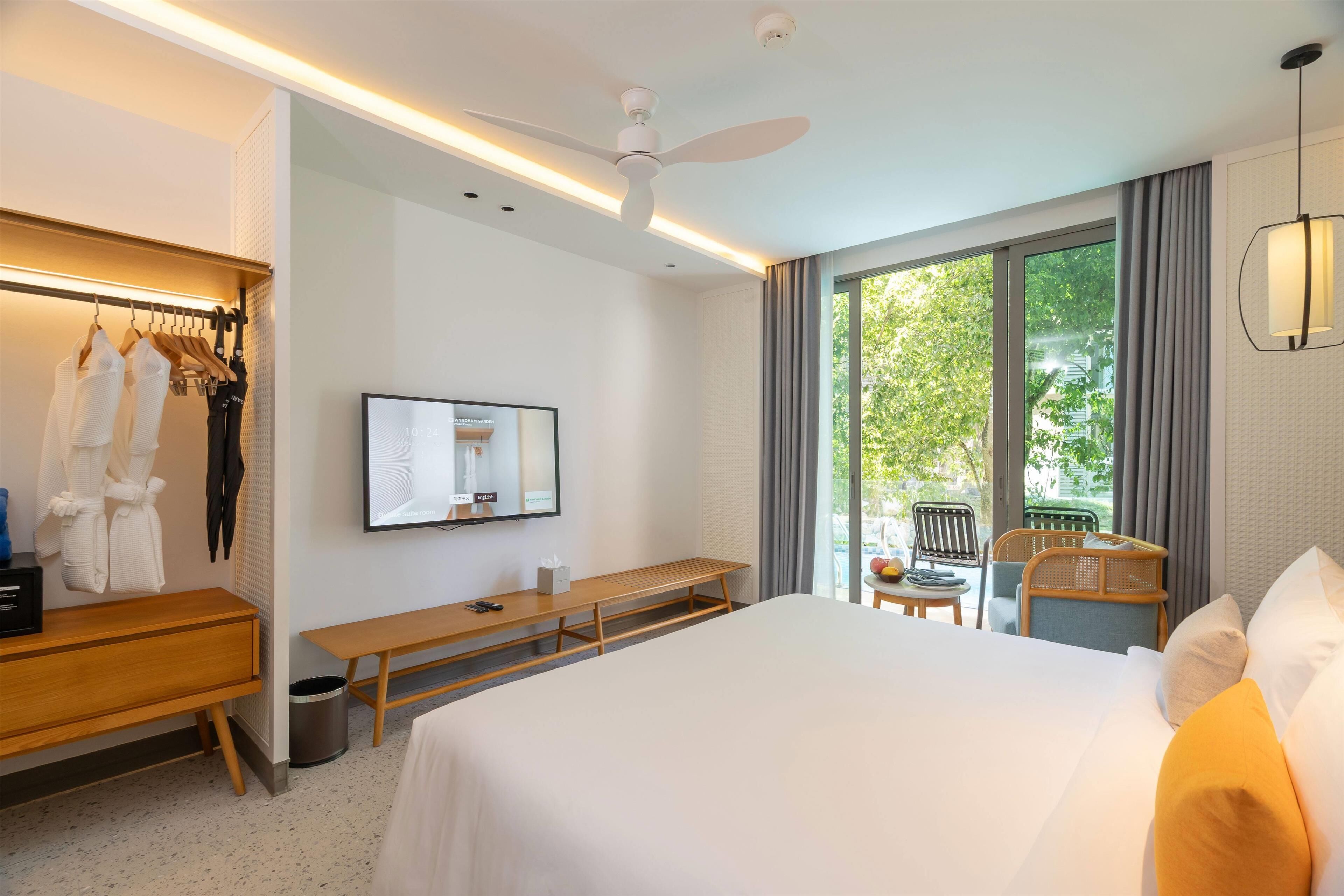 A hotel room with a large bed, a desk, a chair, a TV, and a window with a view of greenery.