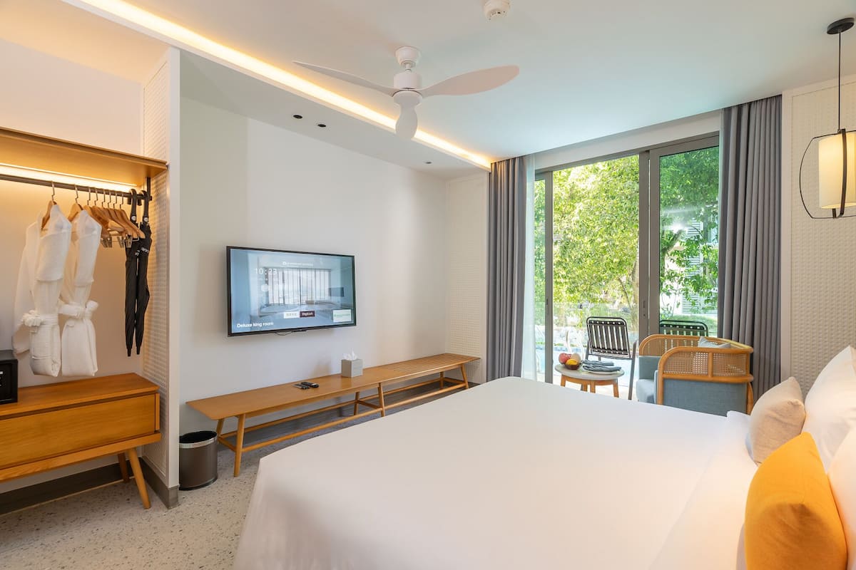 Une chambre d’hôtel équipée d’un lit, d’une télévision, d’un bureau, d’une banquette, d’une chaise et d’une fenêtre donnant sur un espace verdoyant.
