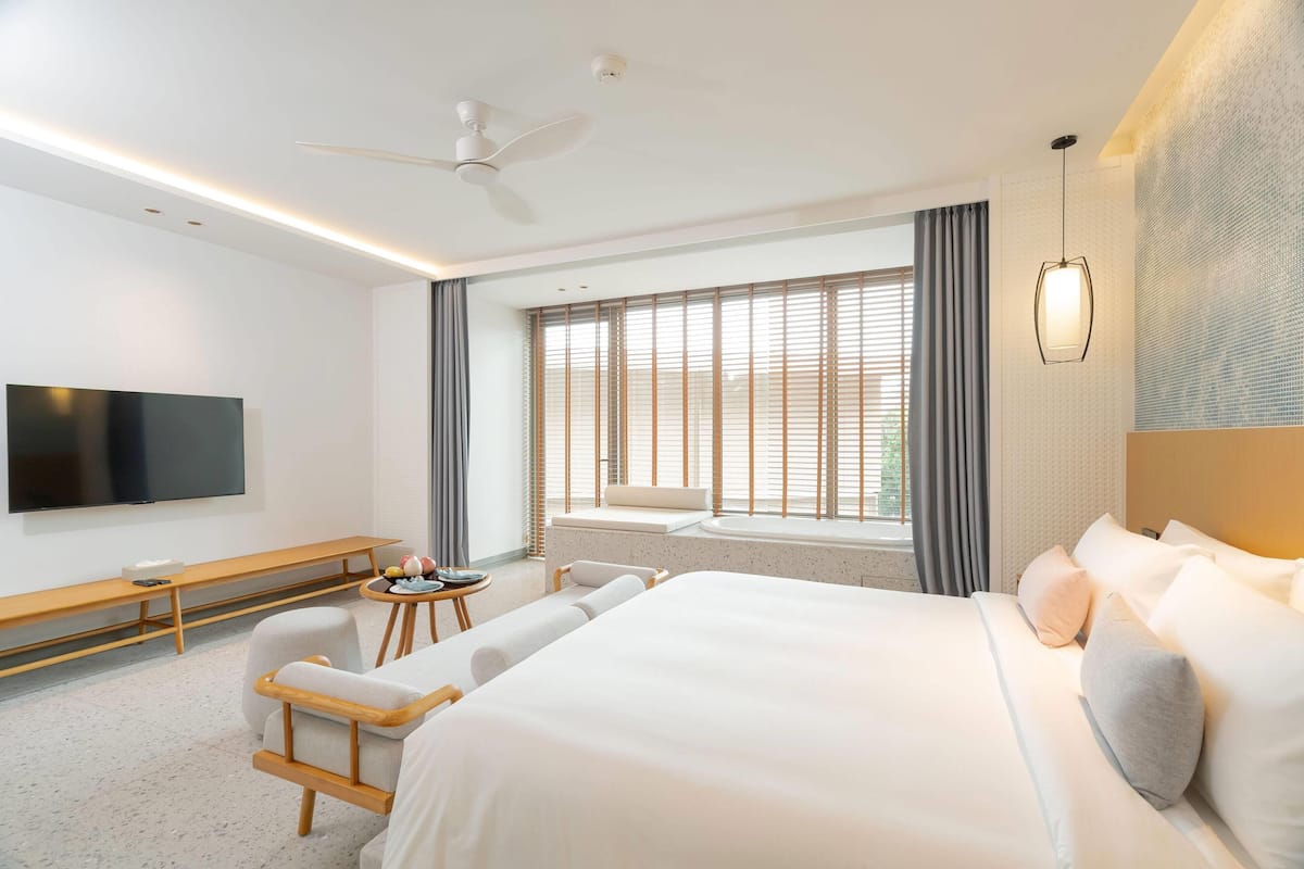 Une chambre d’hôtel moderne dotée d’un grand lit, d’un téléviseur à écran plat, d’un ventilateur de plafond, d’une fenêtre avec stores et d’un petit coin salon avec une table et des chaises.