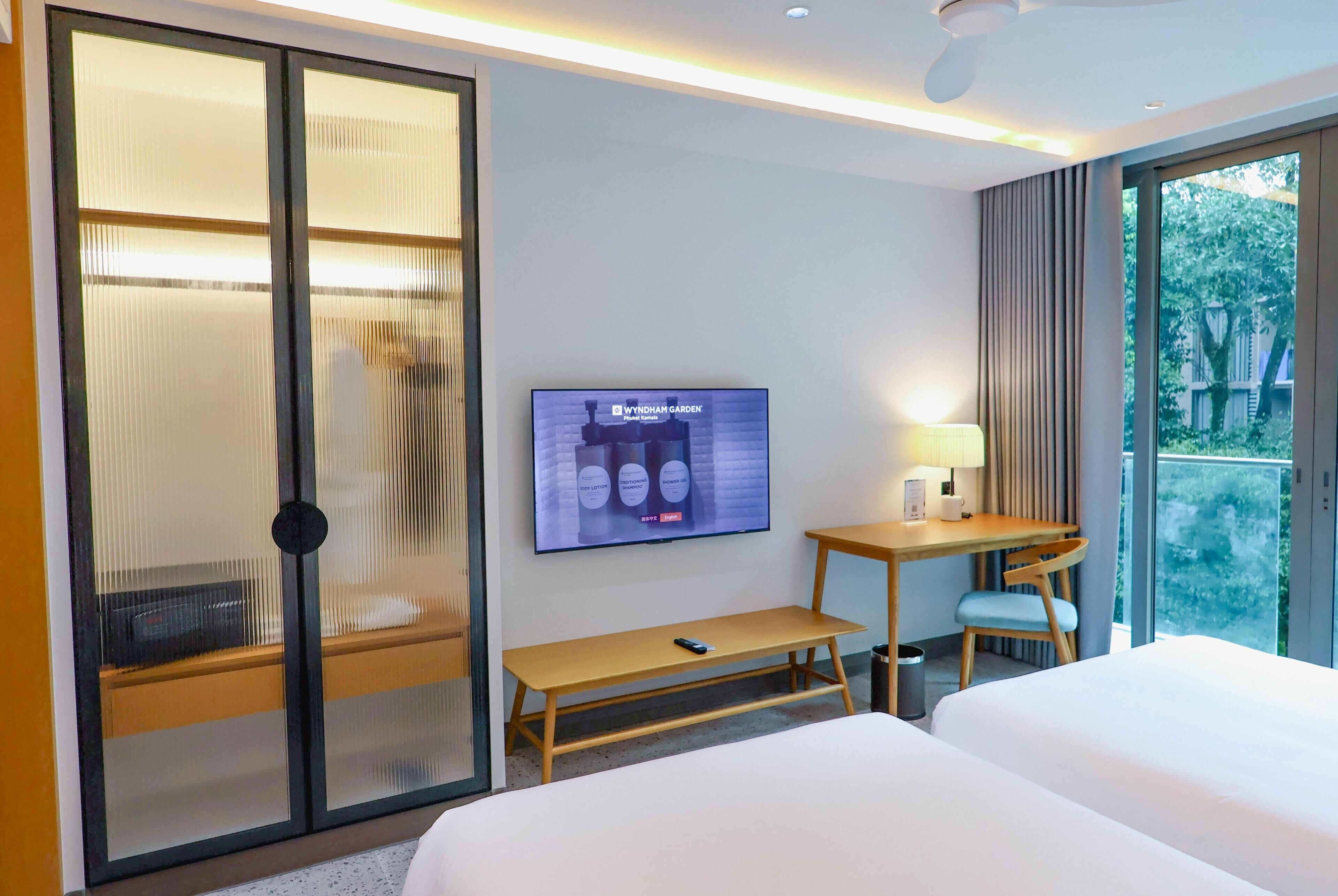 Habitación de hotel con cama, televisor montado en la pared, escritorio con lámpara y vistas al verdor exterior.