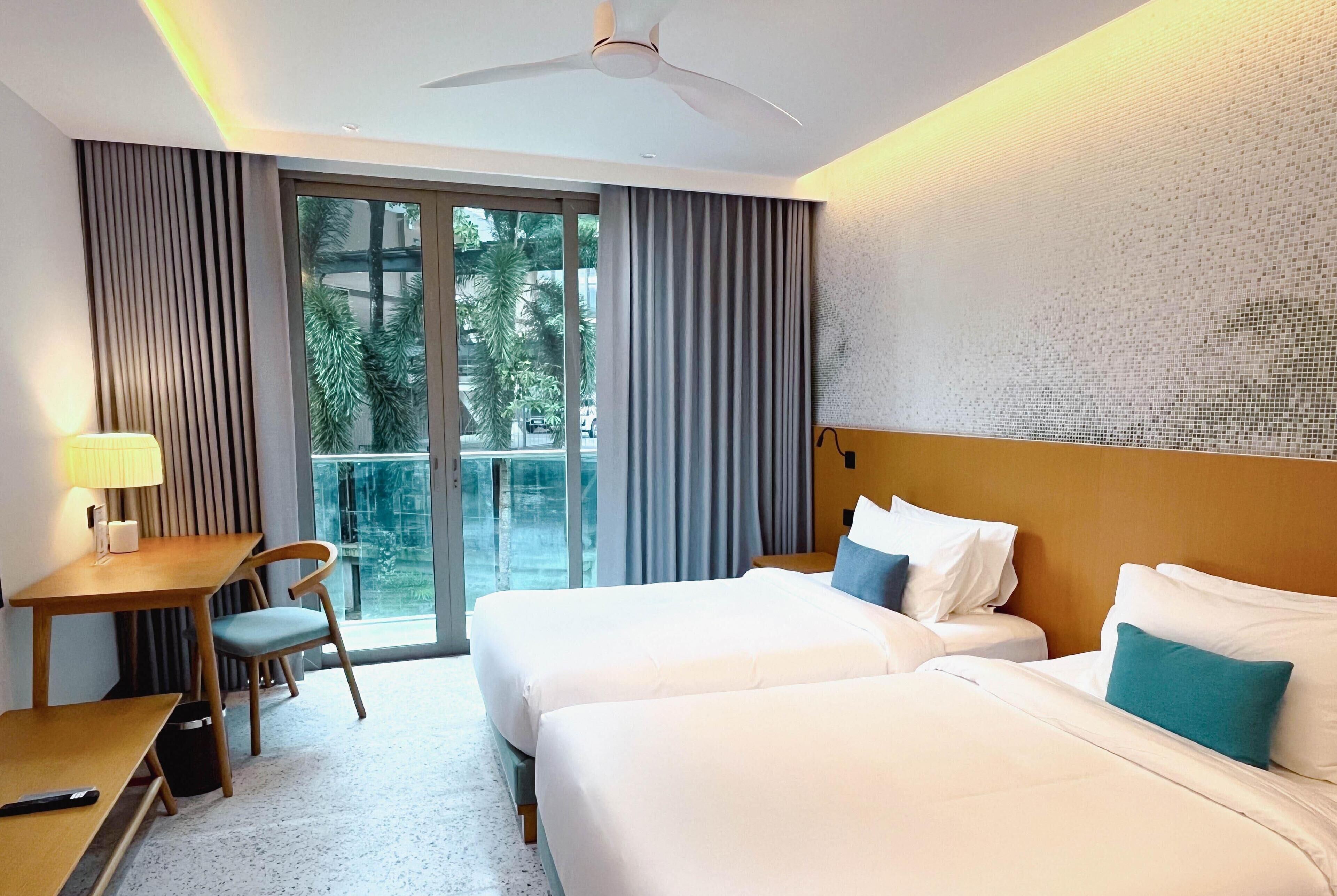 A hotel room with two beds, a desk, a chair, and a view of greenery through the window.