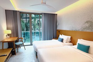A hotel room with two beds, a desk, a chair, and a view of greenery through the window.