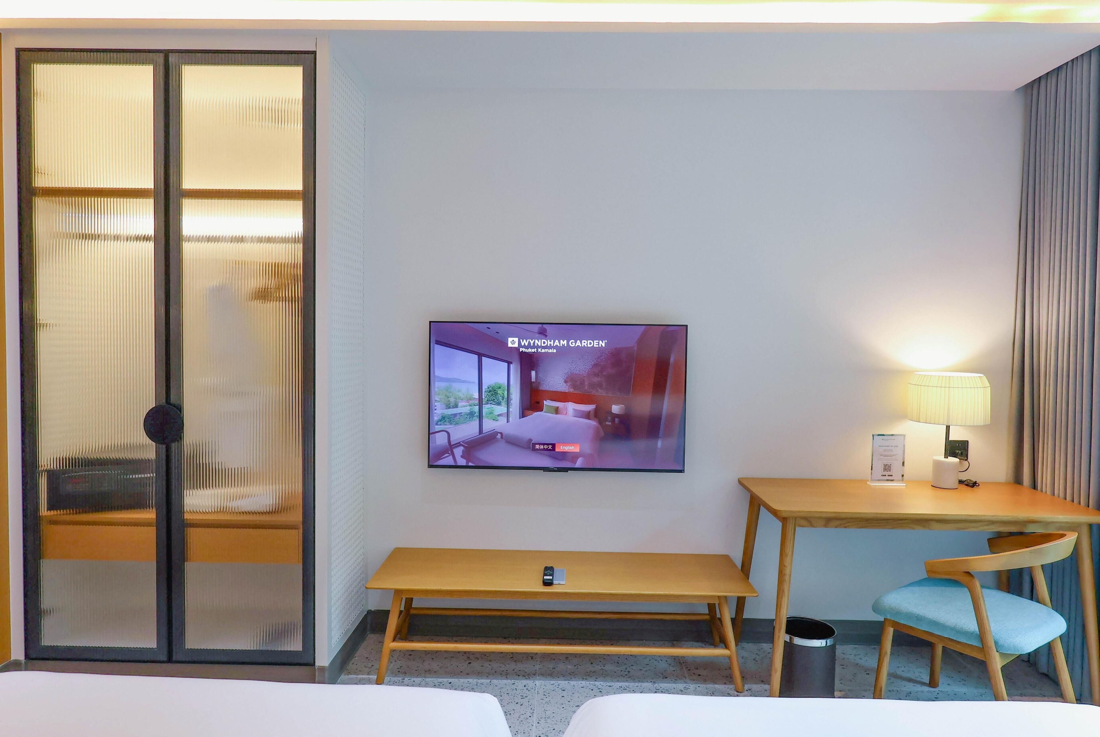 A hotel room with a bed, a TV on the wall, a desk with a chair, and a sliding door with blinds.