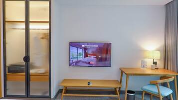 A hotel room with a bed, a TV on the wall, a desk with a chair, and a sliding door with blinds.