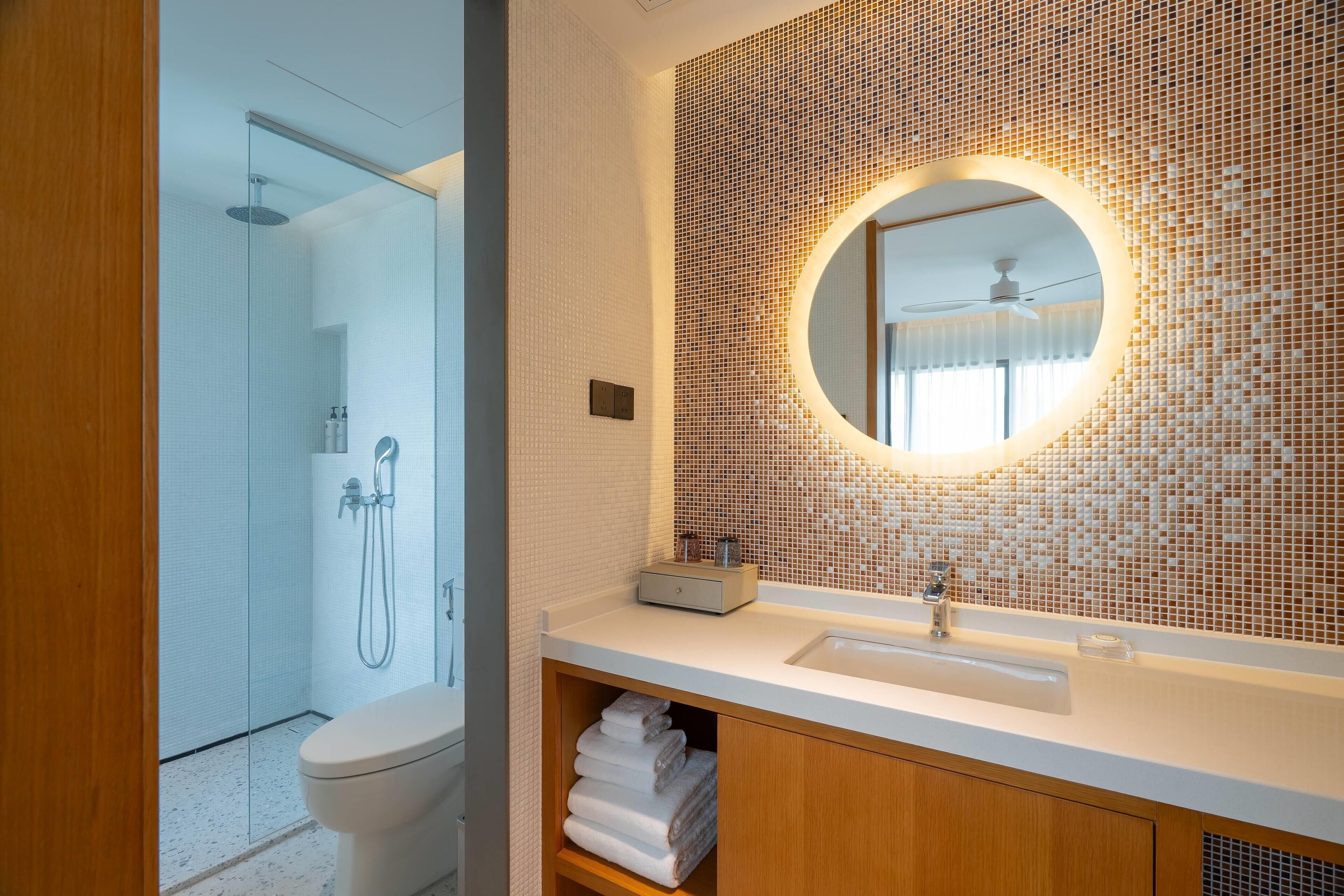 A modern bathroom with a glass-enclosed shower, a round mirror with a mosaic backsplash, and a wooden vanity with a sink.