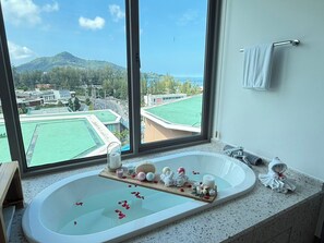A bathroom with a large bathtub, a wooden tray with bath items, and a view of a green field and buildings.
