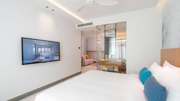 Ein modernes Hotelzimmer mit einem großen Bett, einem Flachbildfernseher, einem Deckenventilator und einer Glaswand, die in einen Wohnbereich mit einer Couch und einem Esstisch führt.