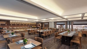 A modern dining area with multiple tables and chairs, a kitchen in the background, and large windows providing natural light.
