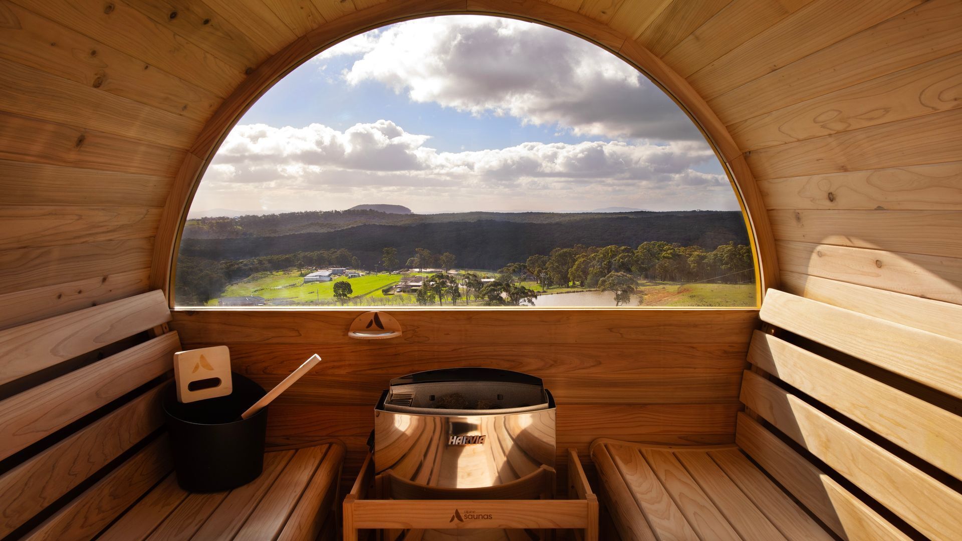 A sauna with a wooden interior, a view of a landscape through a semi-circular window, and a sauna heater.