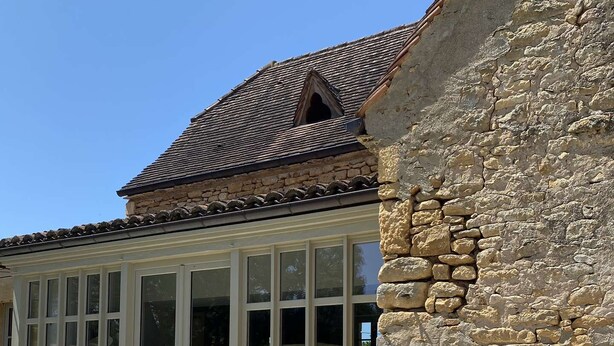 A stone building with a large windowed room, a stone wall, and a metal grill table.