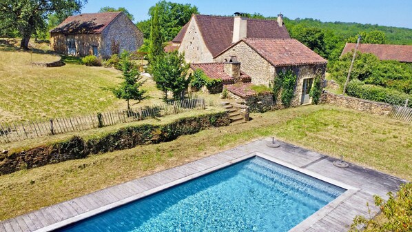 A swimming pool with a wooden deck, surrounded by stone houses with red-tiled roofs, set in a grassy landscape.