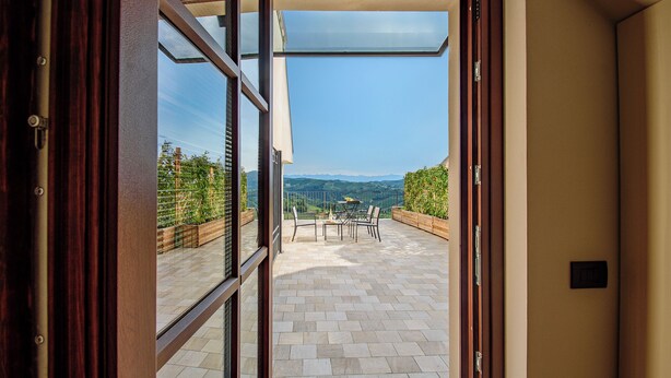 A view through a glass door to a patio with a table and chairs, a wooden fence, and a mountainous landscape. - Elegant Panoramic Two-Room Apartment. Treiso Belvedere 1, Elegante Bilocale Panoramico. Treiso Belve... (Treiso)