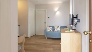 A room with a wooden floor, a blue sofa with floral cushions, a wooden cabinet, and a television mounted on the wall. - Elegant Panoramic Two-Room Apartment. Treiso Belvedere 1, Elegante Bilocale Panoramico. Treiso Belve... (Treiso)