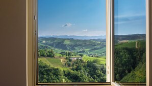 A window with a view of a green valley, a small building, and distant mountains. - Elegant Panoramic Two-room Apartment. Treiso Belvedere Elegance, Elegant Panoramic Two Bedroom Apartment. Treiso Belved... (Treiso)