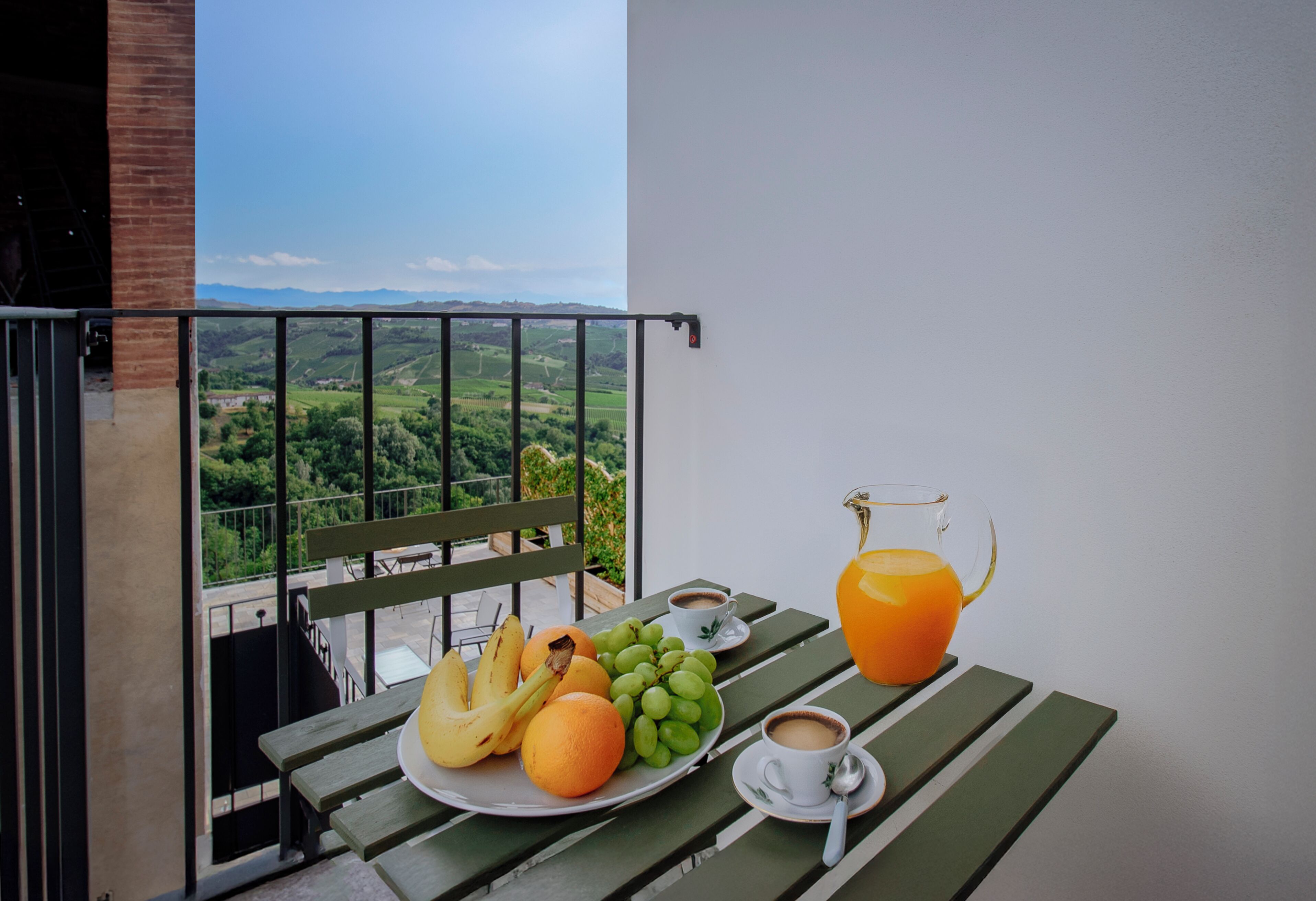 Een balkon met een ontbijttafel, compleet met fruit, koffie en sinaasappelsap, met uitzicht op een prachtig landschap.