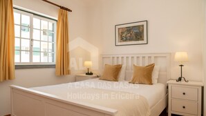 A bedroom with a bed, two bedside tables, a nightstand, a window with curtains, and a framed picture on the wall. - Casa os Naias by ACasaDasCasas (Ericeira)