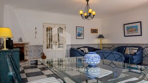 A living room with a glass coffee table, blue sofa, patterned tiles, and a chandelier. - Casa os Naias by ACasaDasCasas (Ericeira)